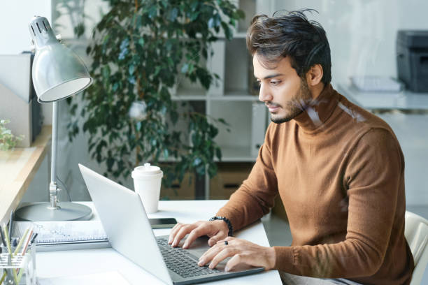 istock Concentrated young Middle Eastern man in brown turtleneck sitting at table and using laptop in office 1355894069