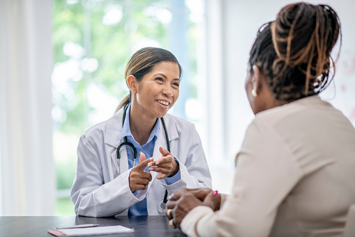 A female doctor meets with a senior patient to discuss her health concerns.  she is seated across her desk from the woman and wearing a white lab coat as she smiles gently.