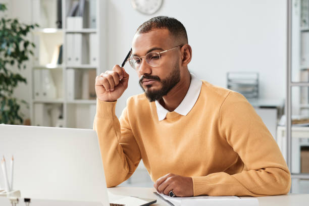 istock Serious busy young middle eastern marketer with beard holding pen and looking at laptop screen while working on strategy in office 1355896887