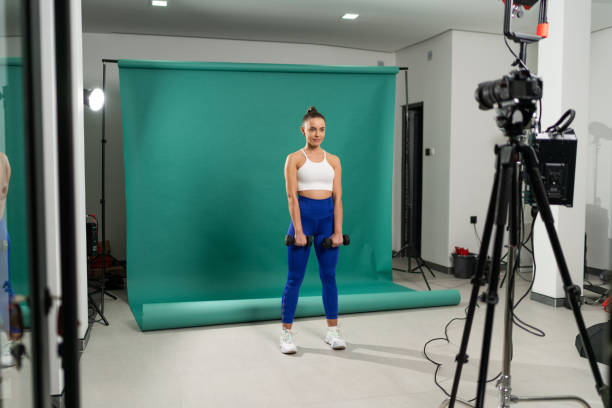 Young female instructor exercising in studio with green background with dumbbell in front of camera