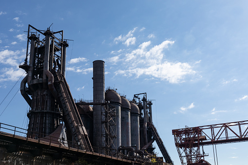 Rows of blast furnaces in an old steel mill silhouetted against a blue sky with clouds, industrial facility shapes, horizontal aspect