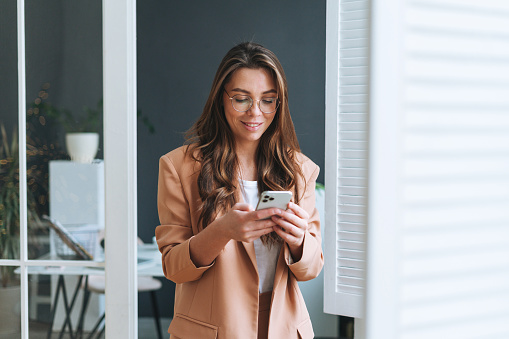Young smiling brunette business woman in glasses with long hair in stylish beige suit using smartphone in modern office