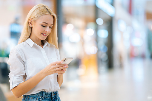 Portrait of smiling attractive blonde young woman with long hair in stylish clothing using mobile phone, standing in hall of mall centre, blurred background, looking at smartphone screen.