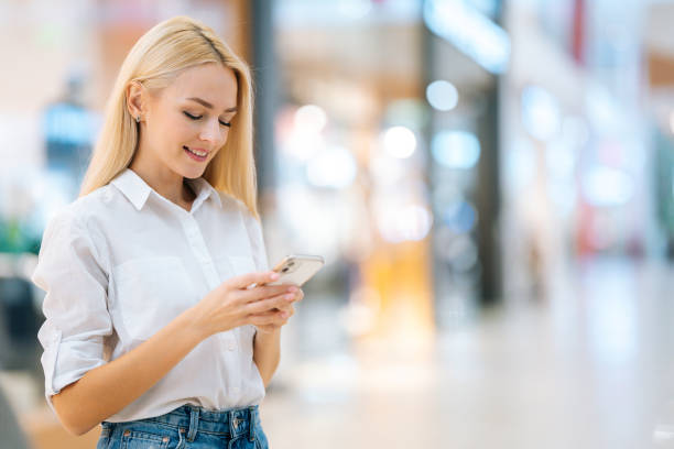 portrait of smiling attractive blonde young woman with long hair in stylish clothing using mobile phone, standing in hall of mall centre, - telephone indoors retail shopping mall imagens e fotografias de stock