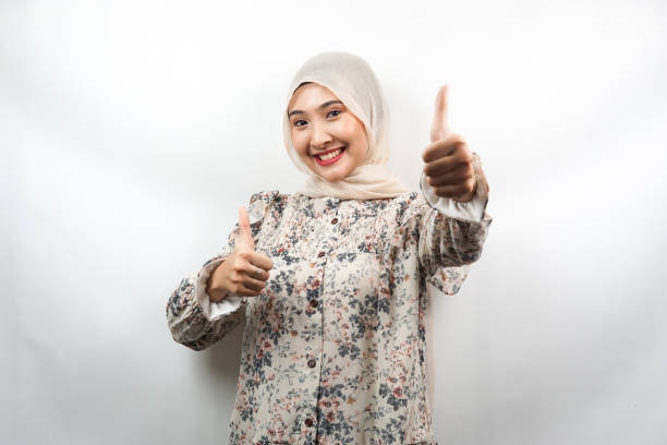 Beautiful young asian muslim woman smiling confident, enthusiastic and cheerful with  hands thumbs up, ok sign, success, good job, success sign, isolated on white background stock photo