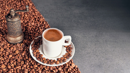 Top-view of a Turkish coffee in a white cup and a coffee grinder. There are coffee beans on the saucer of the coffee cup. The coffee cup and the grinder are on coffee beans. There are coffee beans on the ground but they are cut in half. One half has coffee beans the other half doesn’t.