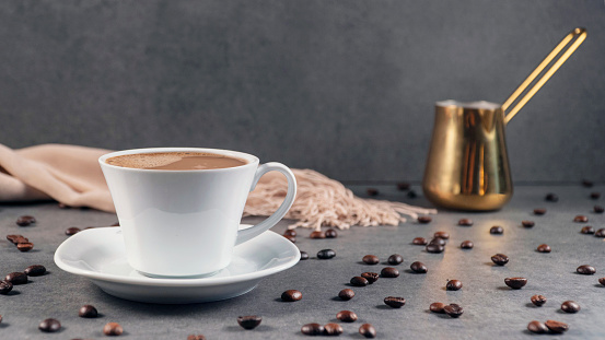 The photo is about a set of rather elegant coffee cups. The photo is mainly consisting of items related to coffee. Raw coffee beans are scatting around on a gray blank ground. In the background, there is also a metal goldish coffee pot which looks quite expensive and charming. The photo makes one feel the freshness and creamy form of the coffee. The background of the photo, except for the items, is gray and blank. This is a coffee house, coffee making, and espresso concept.
