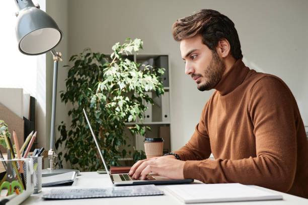 istock Confident young middle eastern manager in sweater typing on laptop while working in office 1355954180