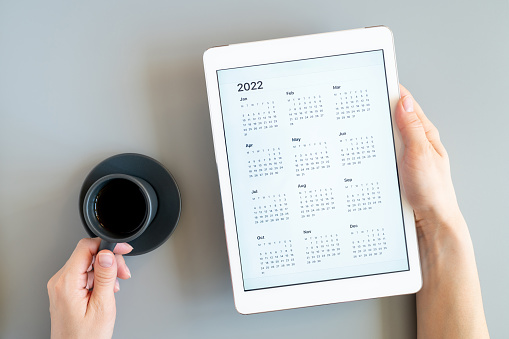 tableta con aplicación abierta de calendario para el año 2022 en manos de una mujer y taza de té o café sobre un fondo gris. concepto de negocio o hacer lista de objetivos con tecnología utilizando. vista superior, lay plano photo