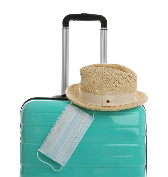 istock Stylish turquoise suitcase, hat and protective mask on white background. Travelling during coronavirus pandemic 1355964711