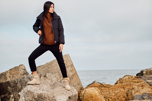 Attractive casual Asian girl in down jacket thoughtfully looking posing away on big stones by the sea