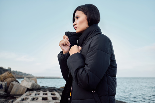 Side view of gorgeous Asian brunette girl in down jacket intently looking away on seaside