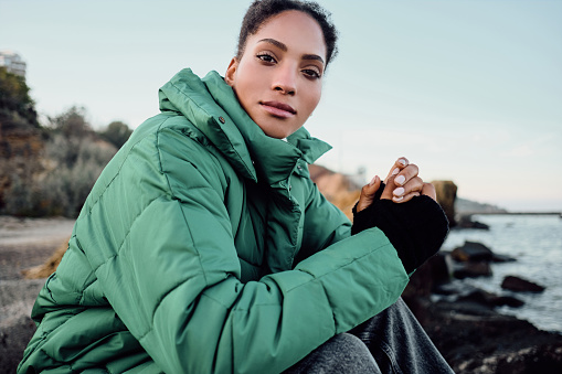Beautiful casual African American girl in down jacket confidently looking in camera sitting on stones by the sea