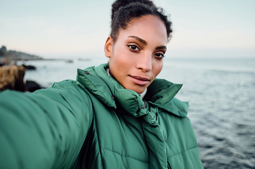 Beautiful casual African American girl in down jacket taking selfie by the sea