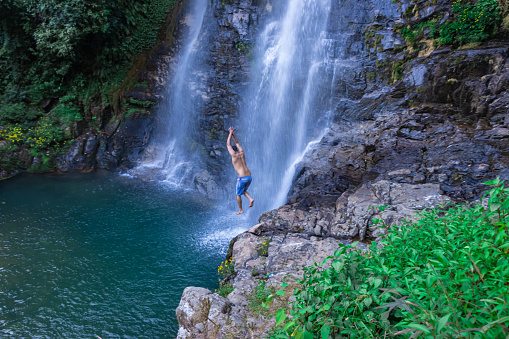 young man jumping from cliff at natural waterfall blue water at morning from top angle image is taken at thangsning fall shillong meghalaya india.