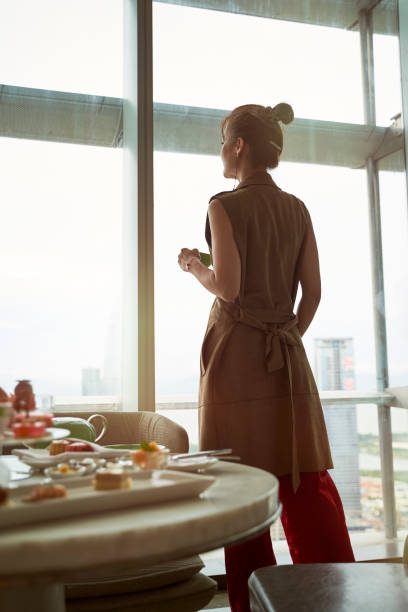 mature asian woman standing in front of window looking at view in hotel room stock photo