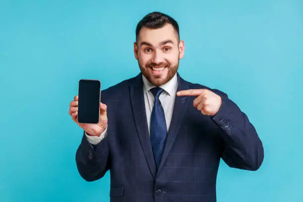 Happy cheerful beaded man in dark suit pointing finger at smartphone with empty screen looking at camera with toothy smile, freespace for adv. Indoor studio shot isolated on blue background.