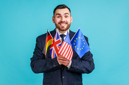 Positive man with beard wearing official style suit holding flags of different countries, language learning, looking at camera with toothy smile. Indoor studio shot isolated on blue background.