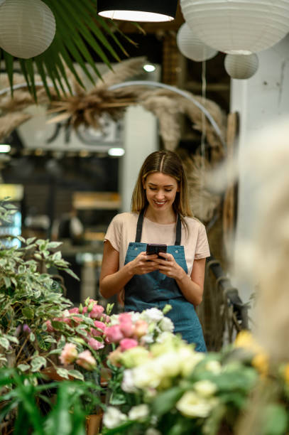 istock Florist using smartphone while surrounded with flowers and plants in a flower shop 1356010191