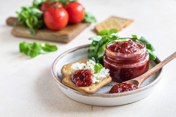 mermelada de tomate, confitura o salsa en tarro de cristal con galletas saladas y ensalada de hojas verdes. mermelada salada inusual. cocina mediterránea. - chutney jar tomato preserved fotografías e imágenes de stock