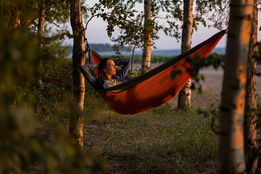 a European woman in a hammock in nature is resting and watching the sunset. a female travel blogger creates video content. hiking and outdoor recreation by the sea or in the woods. a cozy hammock among the trees and a happy millennial woman. silhouette of