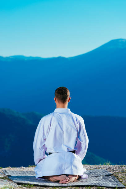 istock Karate master meditating on top of a mountain wearing kimono while looking down at the mountain lake. 1356017020