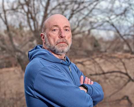 harsh light, casual, backyard portrait of a senior, confident, serious, bald man in sweatshirt with trees in background