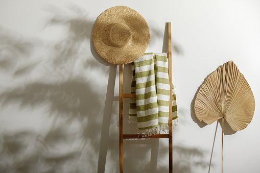Organic linen beach towel on wood ladder, straw hat and dried palm leaf against white wall with shadows from tree leafes
