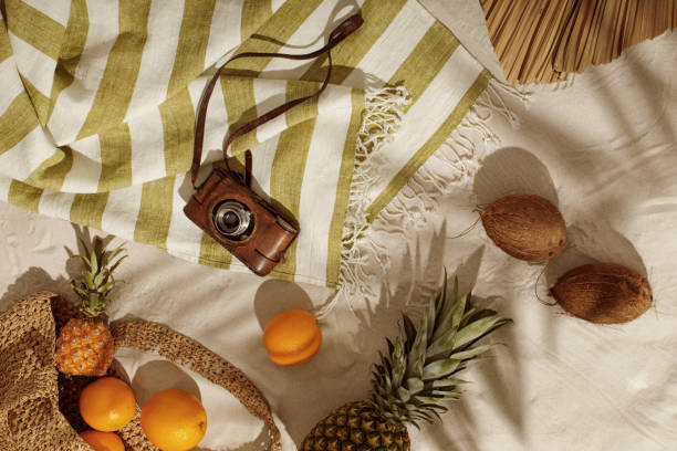 istock Fresh oranges, pineapple, coconuts falling out from woven bag on sandy beach. Vintage camera on linen beach towel with fringes. Tropical holidays concept 1356024912