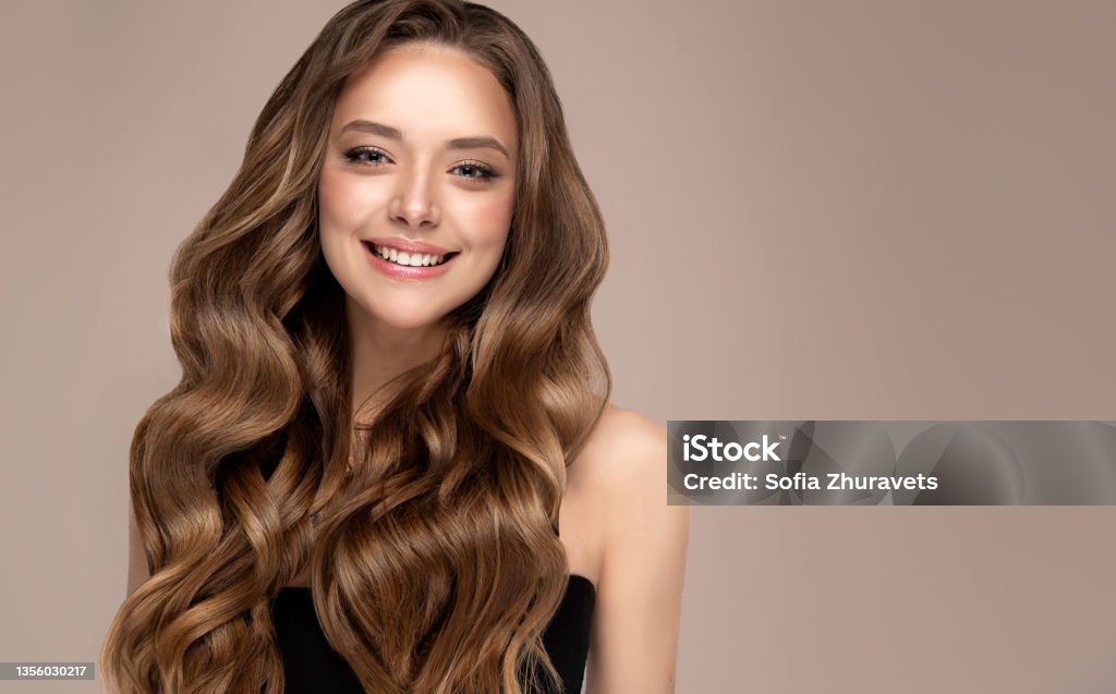 Wide smile on the face of young, brown haired beautiful model with long, well groomed hair. Hair care and hairdressing art. Portrait of beautifully looking young brown haired woman, dressed in a delicate evening makeup.Perfect model straightly gazing by kind, tender look at viewer. Beauty, elegance, hairstyling. Straight Hair Stock Photo