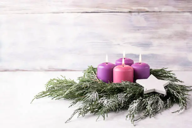 Wreath with four burning purple advent candles on a white wooden background with festive bokeh lights, Christmas catholic tradition.