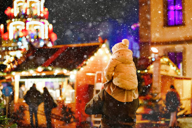 Little preschool girl sitting on shoulder of father on Christmas market in Germany. Happy toddler child and man observing traditional decorated pyramid. Happy family, bonding, love. Family xmas time. stock photo