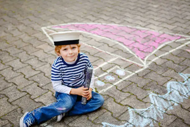 Little kid boy as pirate on ship or sailingboat picture painting with colorful chalks on asphalt. Creative leisure for children outdoors in summer. Child with captain hat and binoculars