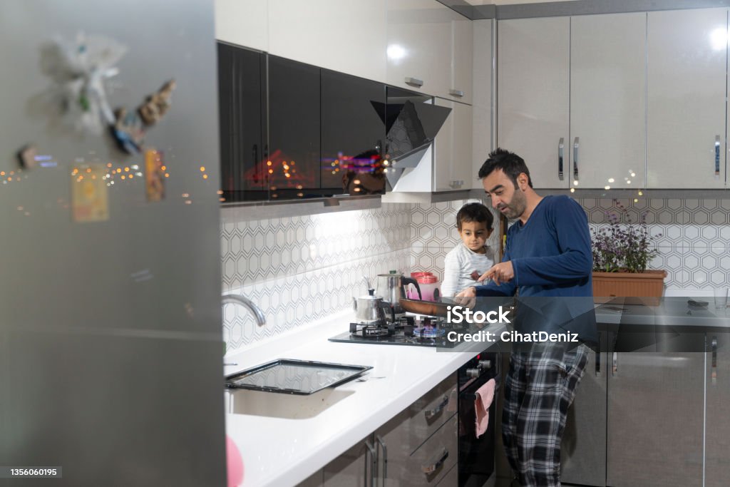 baba oğluna ocakta yiyecek pişiriyor acıkan oğluna gece vakti ocakta yiyecek pişiren baba. pijama giymiş, oğulları onu izliyor.  Mutfak dolaplarından şehir ışıkları yansıyor. Natural Gas Stock Photo