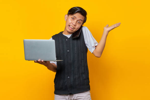 istock Young handsome businessman talking on a smartphone while holding a laptop on yellow background 1356061276