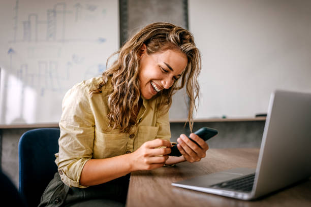 Women using smartphone and laptop laughing Cheerful and beautiful young successful business women sitting in modern coworking space and office laughing while using smartphone and laptop easy stock pictures, royalty-free photos & images