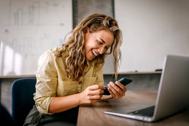 Cheerful and beautiful young successful business women sitting in modern coworking space and office laughing while using smartphone and laptop