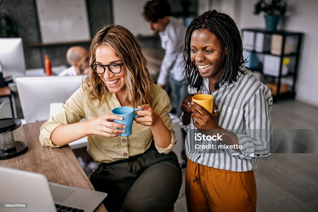 Female coworkers looking at laptop and laughing Female colleagues are commenting and enjoying laptop content at the work in a friendly atmosphere in the office while drinking coffee Employee Stock Photo