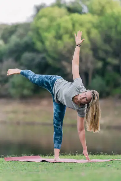 Vertical photo of a woman doing the triangle pose of yoga in a park next to a lake