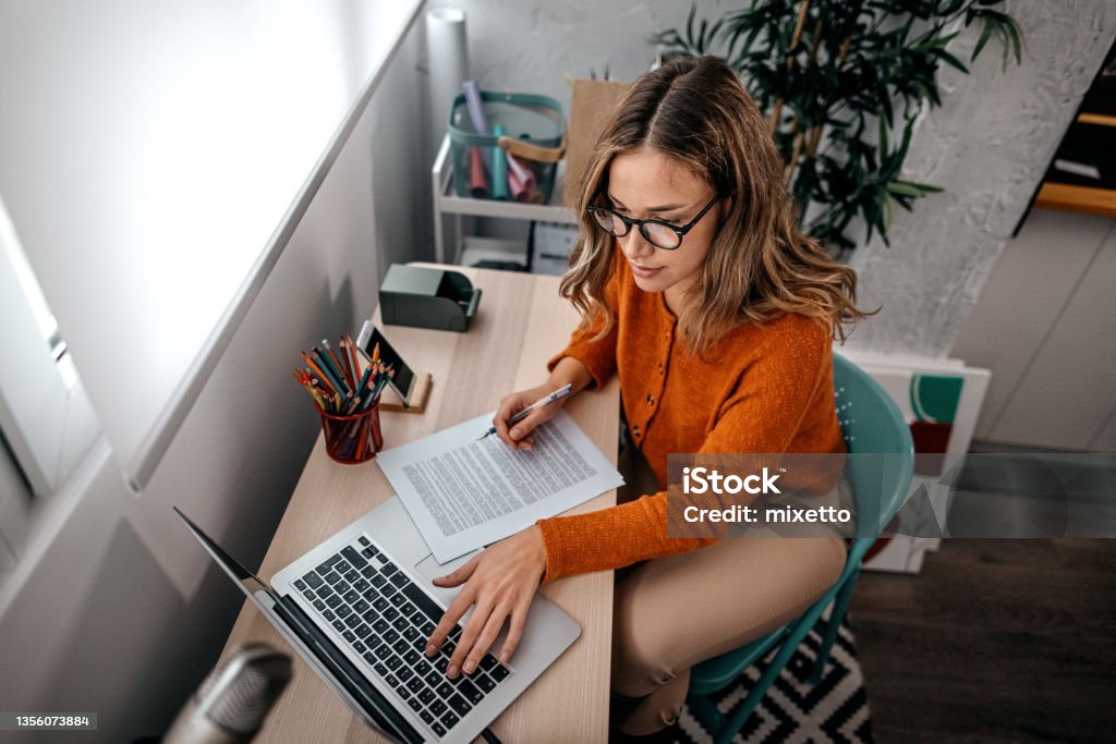 Young woman working on laptop Young woman with spectacles working on laptop while reading with notes at home while podcasting Letter - Document Stock Photo