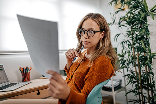 Young woman with eyeglasses thinking and reading document at home office