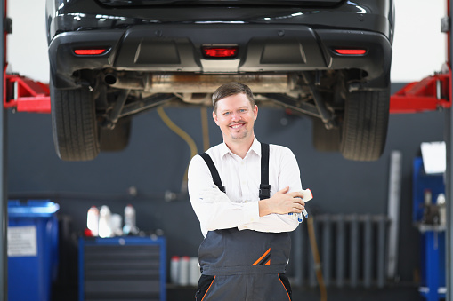 Portrait of qualified mechanic with car behind placed on lifter hoist in garage. Cheerful man like his job. Vehicle service maintenance, pit stop concept