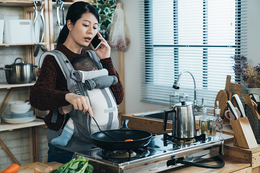 multitasking asian housewife carrying baby with a carrier is speaking on the smartphone while cooking breakfast using frying pan in the kitchen at home.