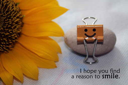 Kindness inspirational quote - i hope you find a reason to smile. Still life with half yellow sunflower and small smiling paper clip on a stone on white background. Kind wishes concept.