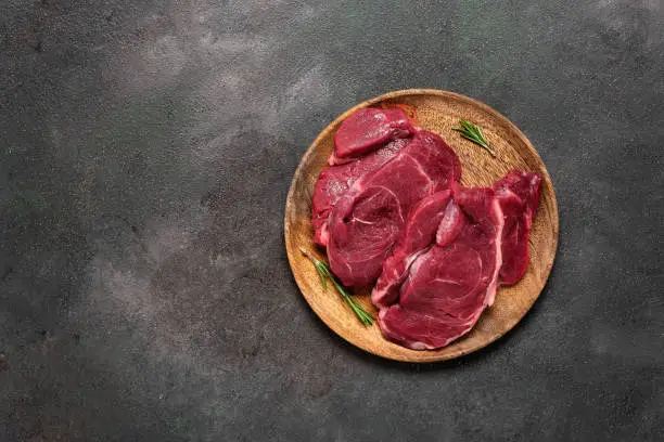 Fresh raw beef steaks on a wooden plate, dark rustic background. Top view, flat lay