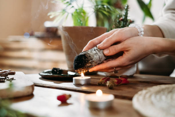 Hands burning white sage for ancient spiritual ritual Woman hands burning white sage, palo santo before ritual on the table with candles and green plants. Smoke of smudging treats pain and stress, clear negative energy traditional ceremony photos stock pictures, royalty-free photos & images