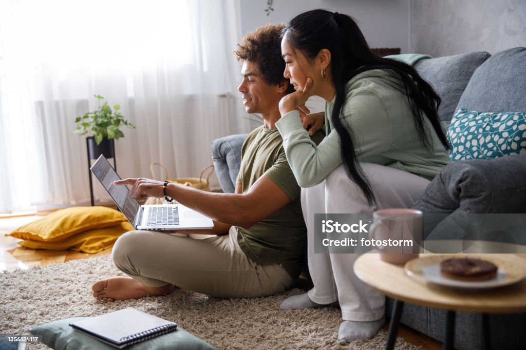 Millennial couple looking at their finances via online banking Wide shot of millennial couple lounging in their living room, planning out their finances and looking at their account via online banking. Home Finances Stock Photo