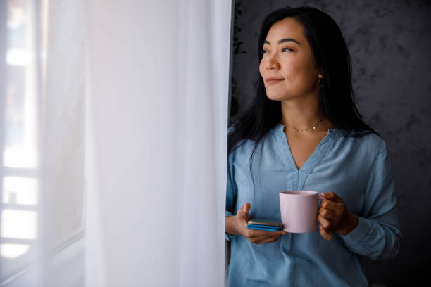 istock Young woman having her morning coffee, using smart phone and looking out the window 1356124698