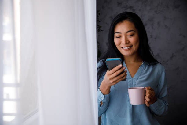 istock Young woman standing by the window, reading a text on smart phone and having coffee 1356124701