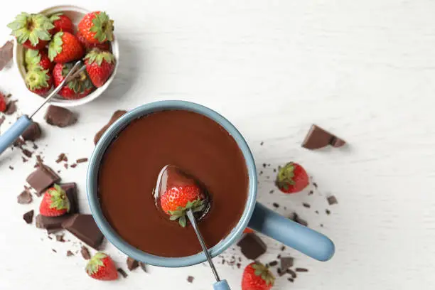 Dipping strawberry into fondue pot with chocolate on white table, top view. Space for text